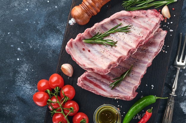 Costolette di maiale crude o carne cruda fresca con spezie su vassoio di legno nero con paprika, spicchi d'aglio ed erbe aromatiche. fondo strutturato scuro con lo spazio della copia per testo.