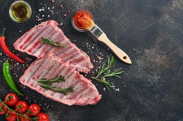 Raw pork ribs or fresh uncooked meat with spices on black wooden tray with paprika, garlic cloves and herbs. Dark textured background with copy space for text.