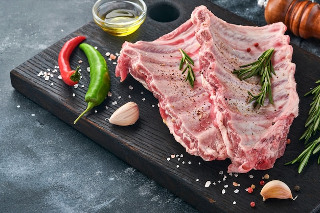 Raw pork ribs or fresh uncooked meat with spices on black wooden tray with paprika, garlic cloves and herbs. Dark textured background with copy space for text.