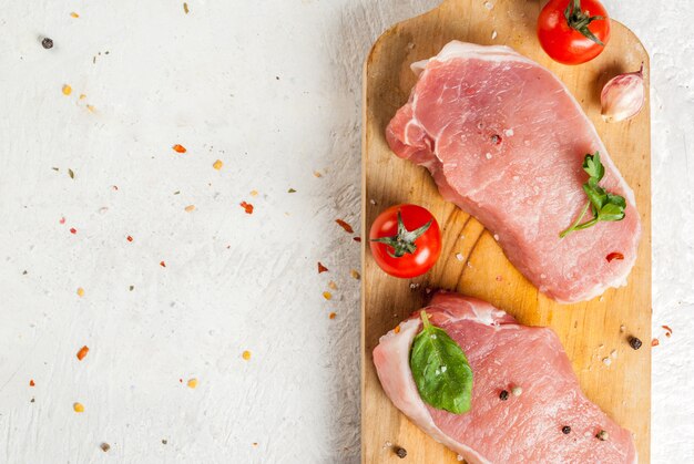 Raw pork, steak, cutlet. Meat, a source of protein. On a cutting board, on a white stone table. With spices, herbs and tomatoes for cooking. Copy space top close view