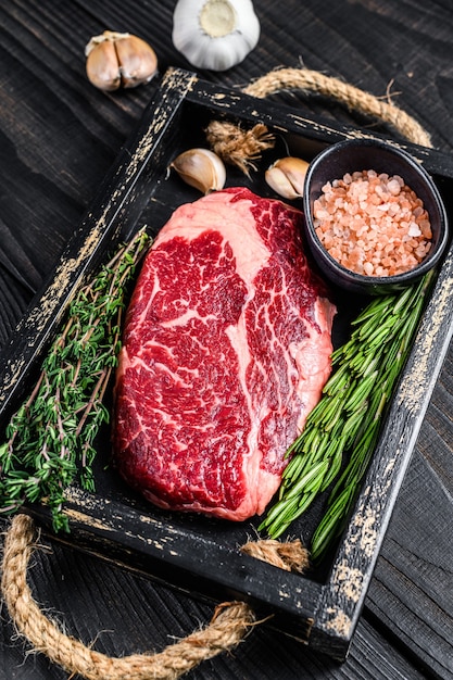 Raw prime rib eye beef meat steak in a butcher wooden tray with herbs. Black wooden background. Top view.