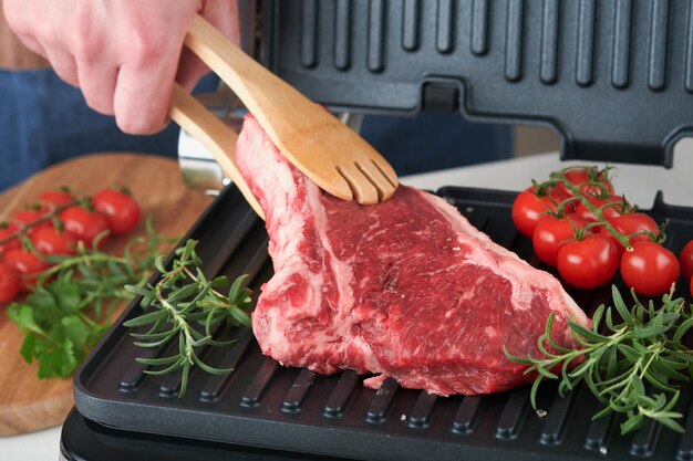 Raw prime steaks on electric grill Chef male hand holding steak Ribeye on modern electric grill with rosemary cherry tomato and pepper Cooking process in modern kitchen Top view copy space