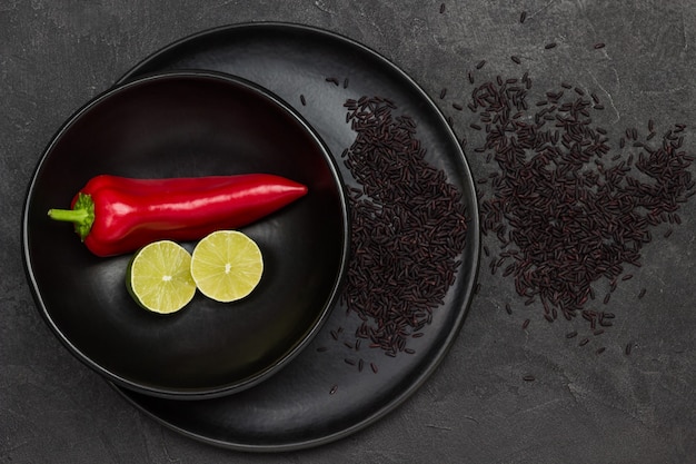 Raw red pepper and lime halves in black bowl. Black rice in black plate. Rice on table. Flat lay. Black surface.