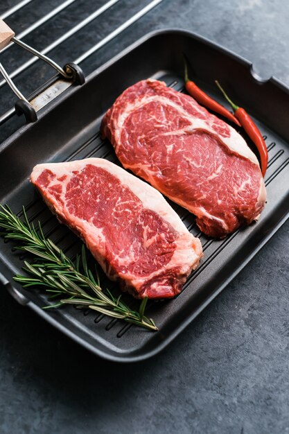 Raw rib eye and striploin New York beef steak on black grilling pan with chili pepper and rosemary