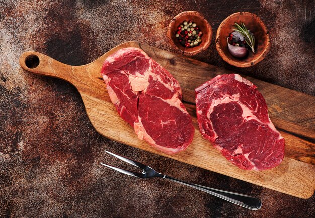 Raw ribeye steaks, spices and meat fork on wooden cutting board on a brown concrete background, top view