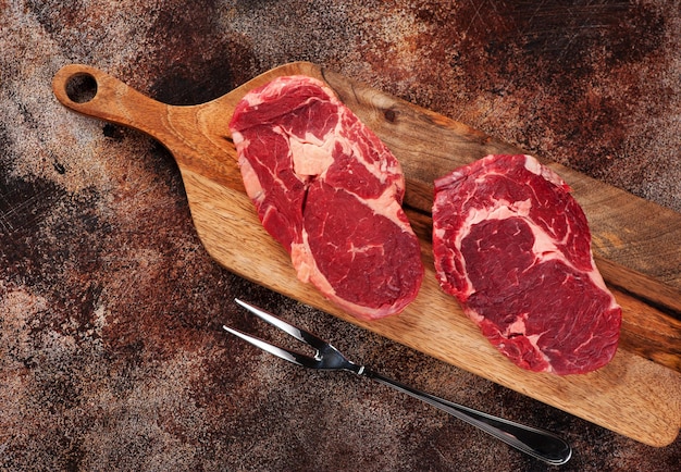 Raw ribeye steaks on a wooden cutting board and meat fork on a brown concrete background, top view