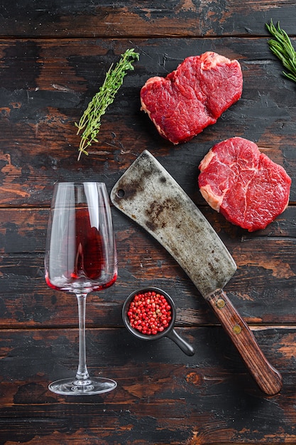 Raw rump steaks with american butcher cleaver and two glass of\
red wine over dark old wooden background, top view.