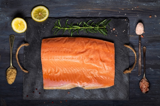 Raw salmon fillet on black slate plate and ingredients for making gravlax.