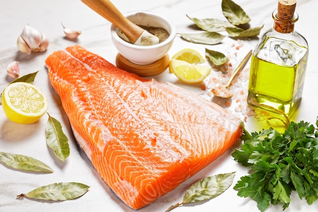 Raw salmon fillet on a marble table with bay leaves, parsley, olive oil, lemon and spices and pink himalayan salt