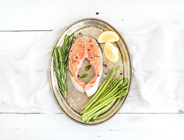 Raw salmon steak with asparagus lemon spices and rosemary on vintage silver tray over white wooden backdrop