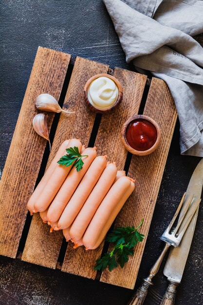 Raw sausages with garlic and sauces on wooden board on dark concrete surface. Top view.