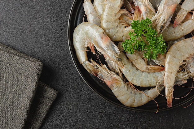 Raw shrimps on black plate. Uncooked seafood with fresh parsley on dark background with napkin. Top view.