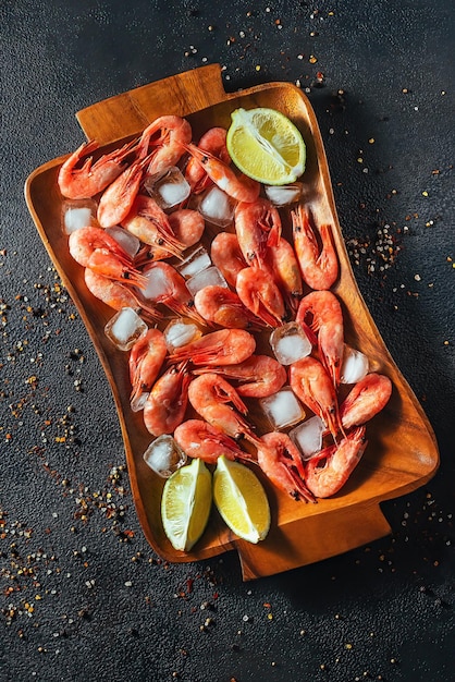 Raw shrimps with ice, lime and rosemary sprigs lie on a wooden tray, dark background