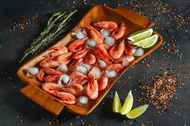 Raw shrimps with ice, lime and rosemary sprigs lie on a wooden tray, dark background.