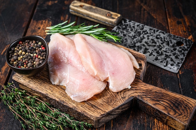 Cotolette di filetto di petto di pollo tagliate a fette crude su un tagliere di legno con mannaia. fondo in legno scuro. vista dall'alto.