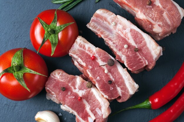 Raw slices of pork on a black slate board, tomatoes, red pepper and powder. Pork belly with