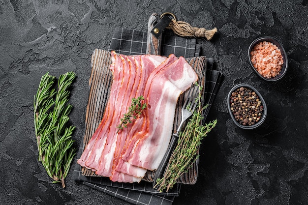 Raw smoked bacon slices with rosemary and thyme on wooden board. Black background. Top view.