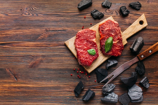 Photo raw steak on cutting board with coal and barbecue fork over wooden textured background