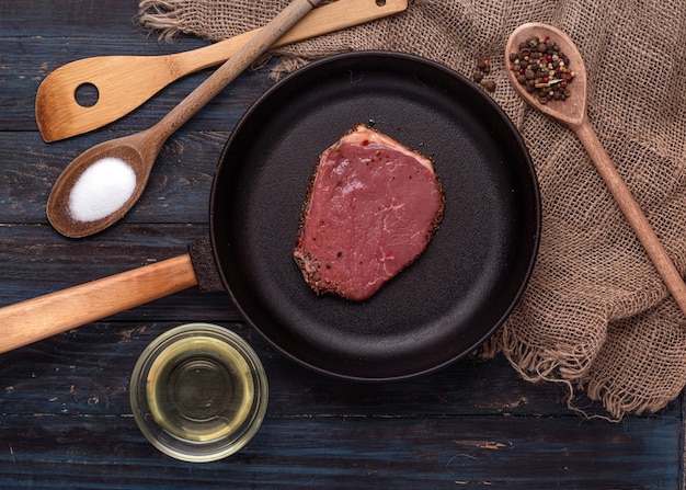 Raw steak in a fry pan, oil, salt and pepper in wooden spoon on wooden background