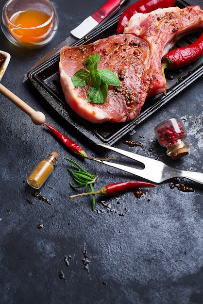 Raw steaks and frying pans with seasoning, garnishes and ingredients on a dark rustic back