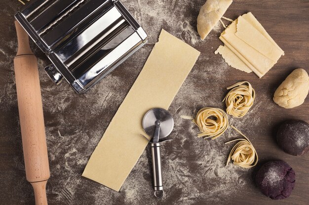 Raw tagliatelle nest and dough slices with wheel cutter on kitchen table. Top view on ingredients and equipment for preparing Italian pasta, copy space