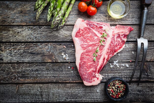 Raw tbone steak with fresh herbs and oil on dark wooden background top view