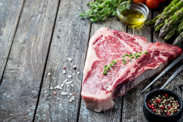 Raw tbone steak with fresh herbs and oil on dark wooden background