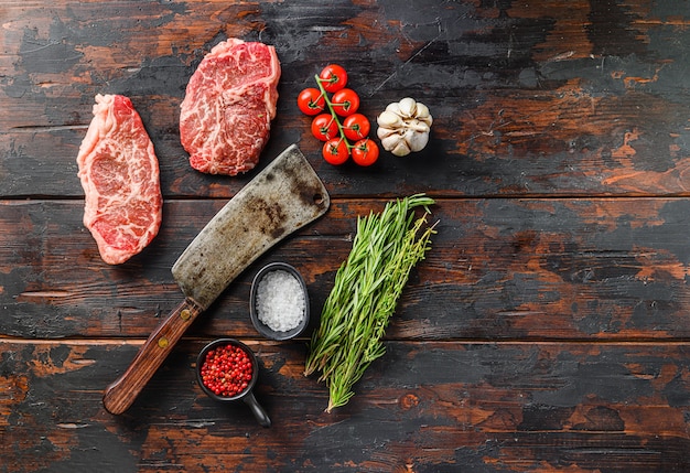 Raw top blade steaks with herbs seasoning and butcher knife over old dark wooden table top view, space for text.