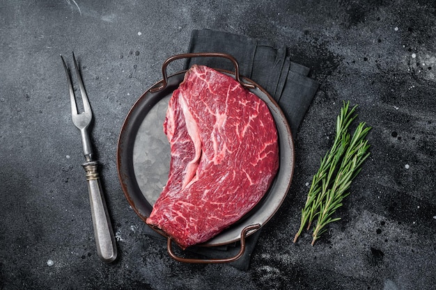 Raw top sirloin beef meat steak in a steel tray with rosemary Black background Top view