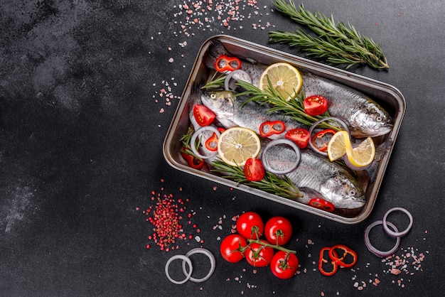 Raw trout lie on a black stone chopping board on a dark concrete table. Nearby are: tomato, rosemary, lemon, pepper, onion, salt