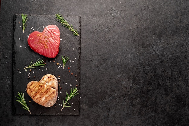 Raw tuna steak on a knife and grilled tuna steak in the shape of a heart with rosemary and spices on a stone background Tuna cooking concept