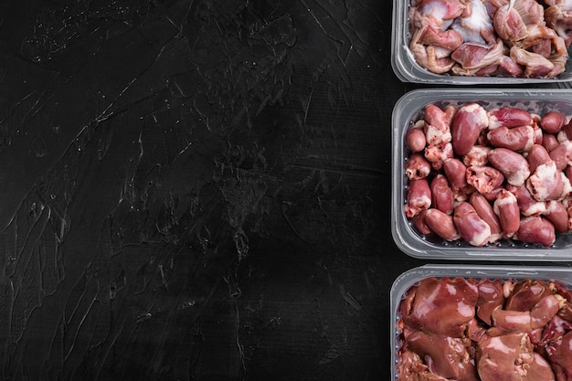 Raw uncooked bird chicken giblets gizzards, stomachs, liver,\
offal, hearts set, on black dark stone table background, top view\
flat lay, with copy space for text