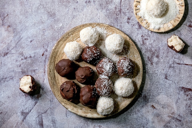 Raw vegan homemade coconut chocolate candy balls with coconut flakes in ceramic plate over white wooden background