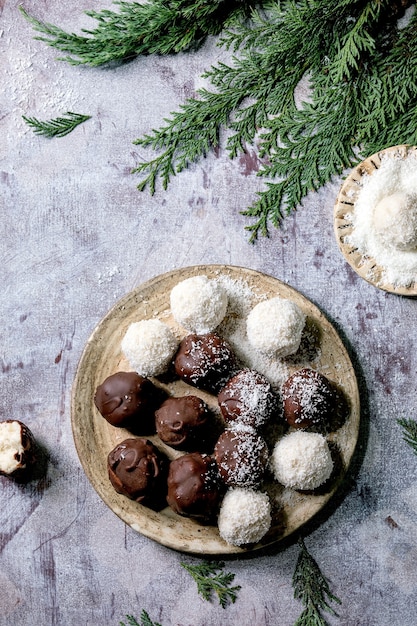 Raw vegan homemade coconut chocolate candy balls with coconut flakes in ceramic plate over white wooden surface. Flat lay, copy space