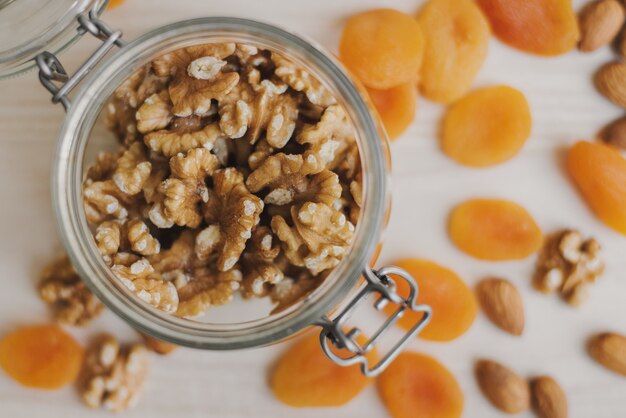 Raw walnut stored in a glass food container and dried apricot and almond on wooden table