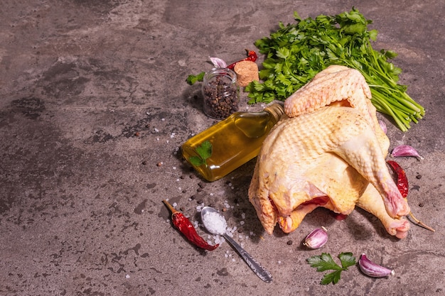 Raw whole chicken carcass for healthy food lifestyle. Free-range farm bird with parsley greens, garlic cloves, spices, and cooking oil. A trendy hard light, dark shadow, stone background, copy space