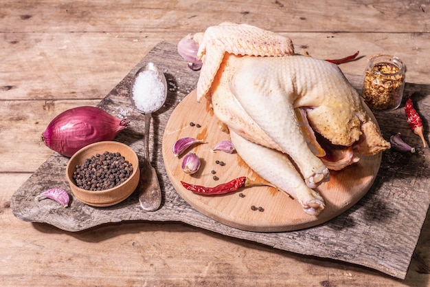 Carcassa di pollo intero crudo per uno stile di vita sano. uccello da fattoria ruspante con cipolla rossa, spicchi d'aglio, spezie e sale marino. una luce dura alla moda, un'ombra scura, un tavolo in legno vintage, spazio per le copie