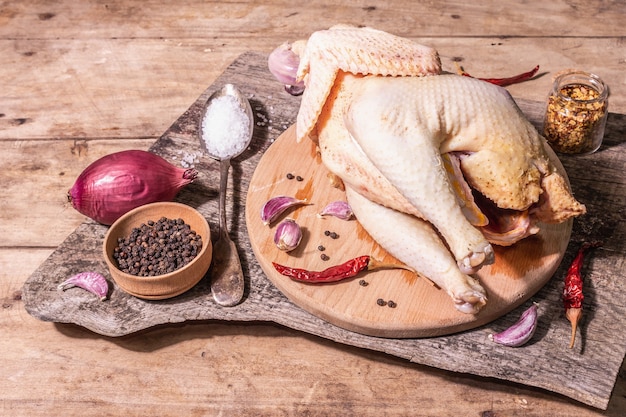 Raw whole chicken carcass for healthy food lifestyle. Free-range farm bird with red onion, garlic cloves, spices, and sea salt. A trendy hard light, dark shadow, vintage wooden table, copy space