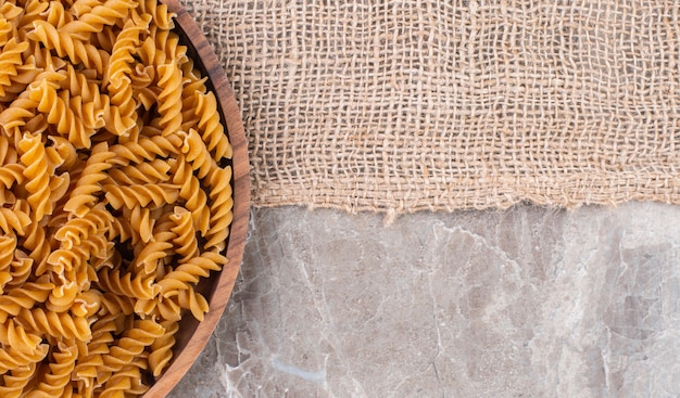 Raw whole wheat fusilli on a wooden plate on a burlap on the marble surface