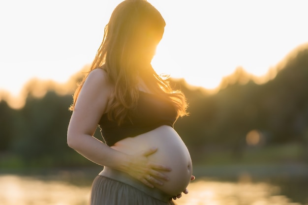 Rays of sunset illuminating a pregnant adult latina woman as she contemplates and touches her belly while standing in a park
