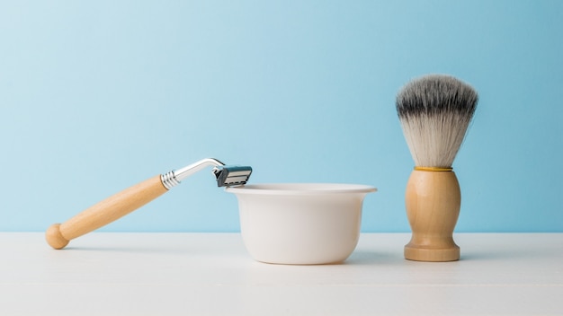 A razor and a brush with wooden handles and a foam bowl on a white table.