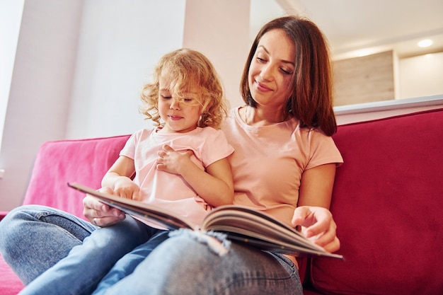 Reading big book Young mother with her little daughter in casual clothes together indoors at home