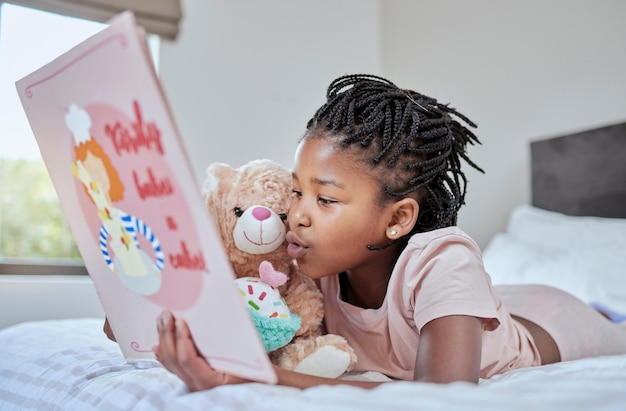 Reading book and teddy bear with a black girl in bed lying down to relax in the bedroom with her hobby children books and education with a female kid telling her teddybear a story in their home