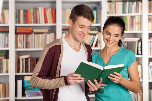 Reading a book together. Cheerful young man and woman reading book together while standing clode to each other in the library