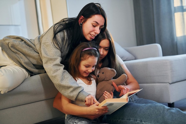 Reading book together Female lesbian couple with little daughter spending time together at home