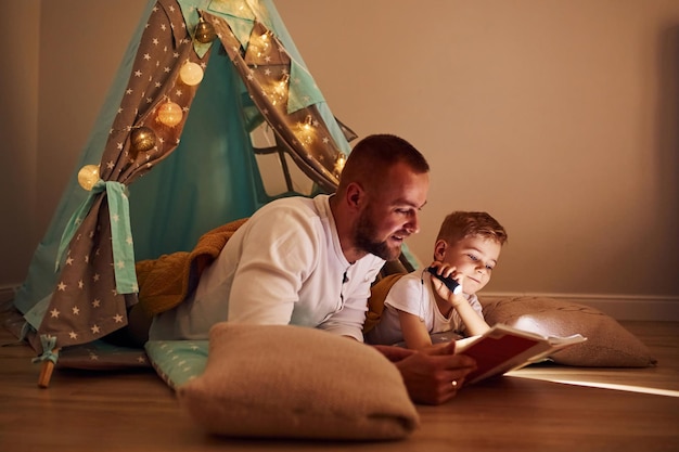 Photo reading book and using flashlight father and his young young spending time together indoors near tent