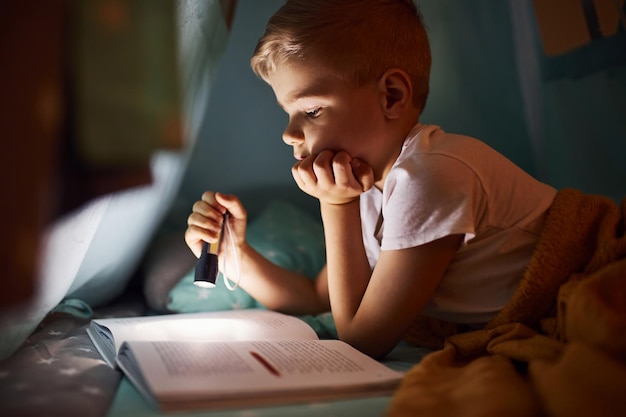 Reading book and using flashlight Young boy in casual clothes lying down near tent at evening time