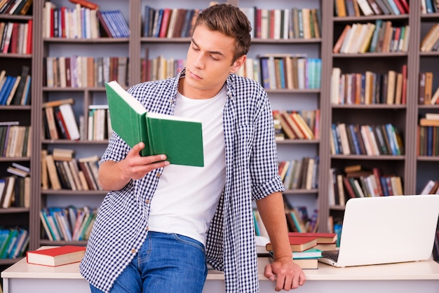 Reading his favorite book. Confident young man reading book while leaning at the desk in library