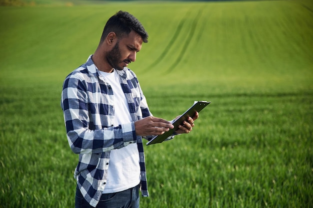 Reading the information from notepad handsome indian man is on the agricultural field