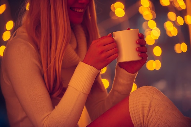 그의 사랑스러운 메시지를 읽고. 흰색 스웨터를 입은 아름다운 젊은 여성이 커피 컵을 들고 배경에 초점이 흐려진 크리스마스 조명을 들고 웃고 있는 모습